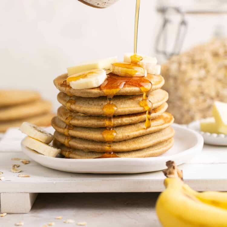 Blender banana oatmeal pancakes stacked up on a plate and topped with banana slices, butter, and maple syrup.