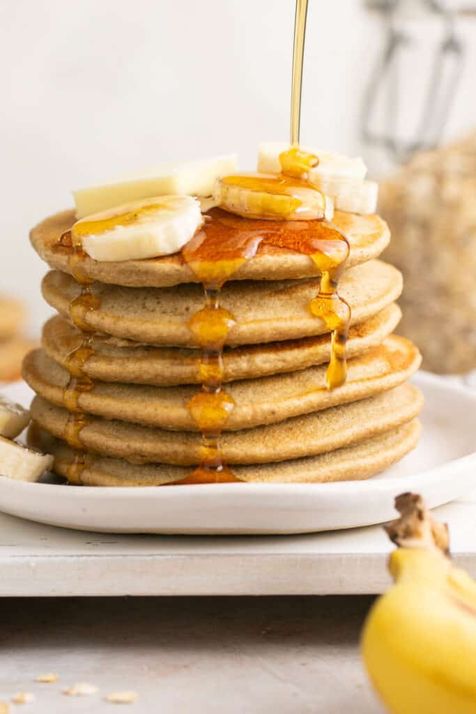 Blender banana oatmeal pancakes in a stack on a plate topped with banana slices and syrup being poured on top.