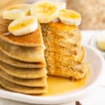 Blender banana oatmeal pancakes in a stack on a plate topped with syrup and banana slices.