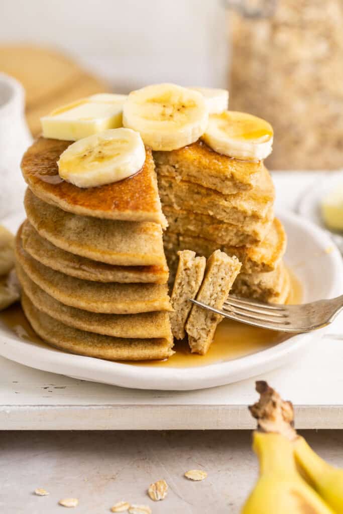 Blender banana oatmeal pancakes in a stack on a plate topped with syrup and banana slices.