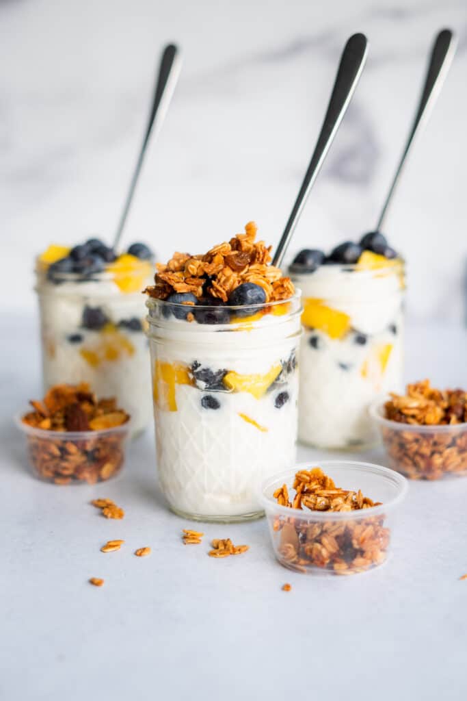 Greek yogurt parfaits with granola and fruit in mason jars with spoons with extra granola in small cups on the side.