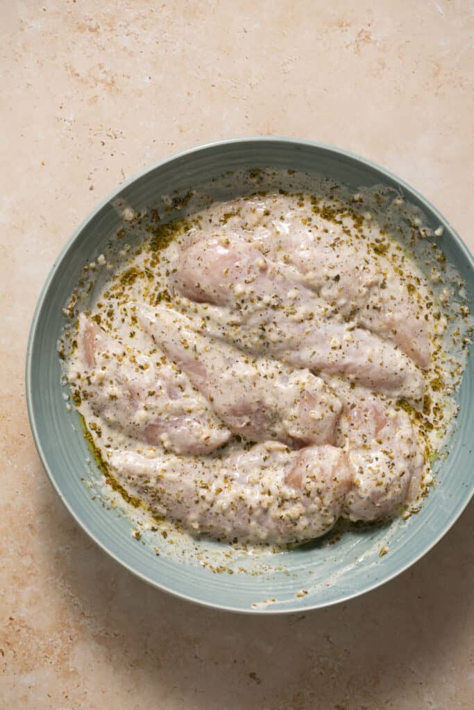 Raw chicken tenderloins covered in the marinade in a bowl.
