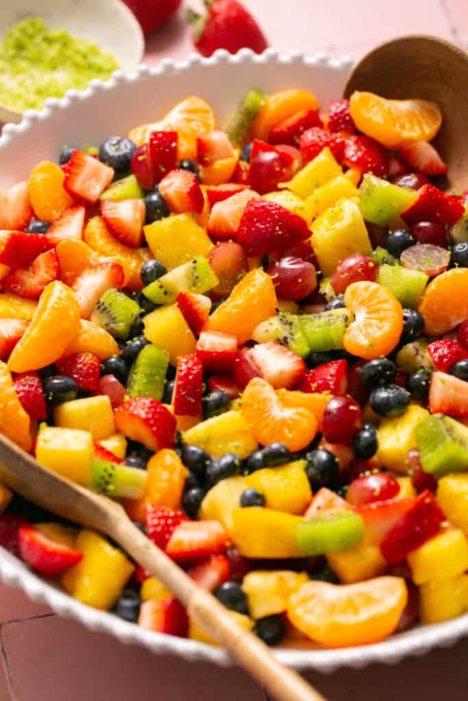 Rainbow fruit salad with honey lime dressing in a large bowl with a wooden spoon.