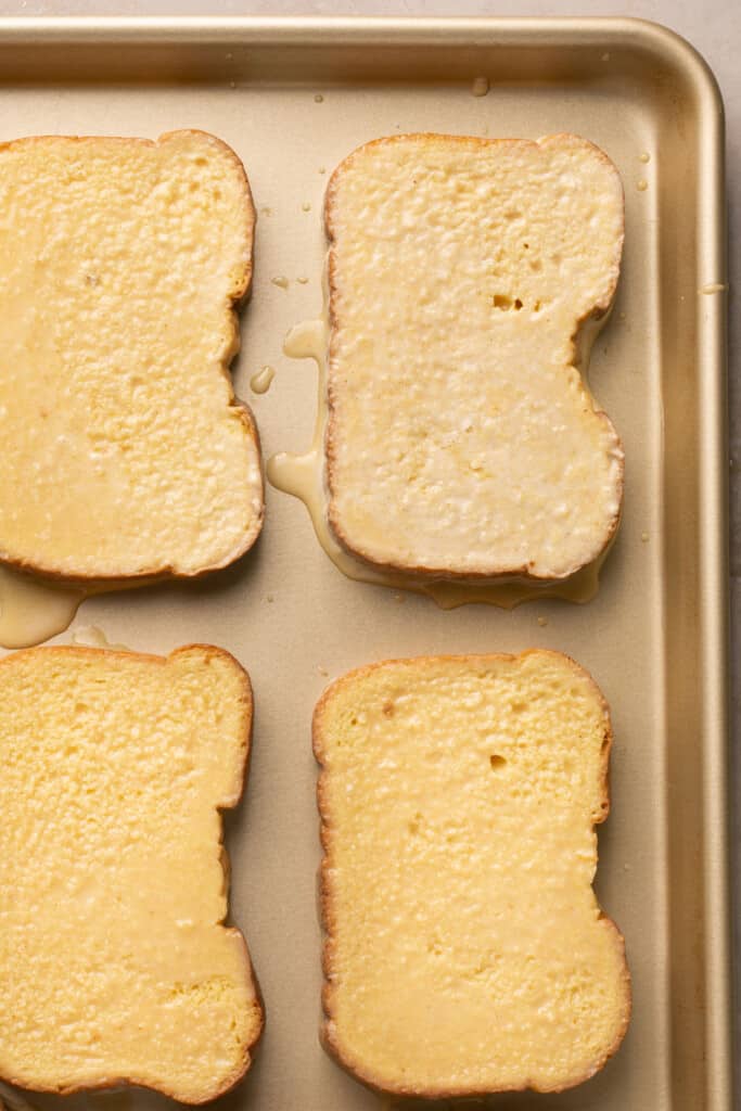 Slices of bread after being dipped in the egg mixture on a baking sheet.