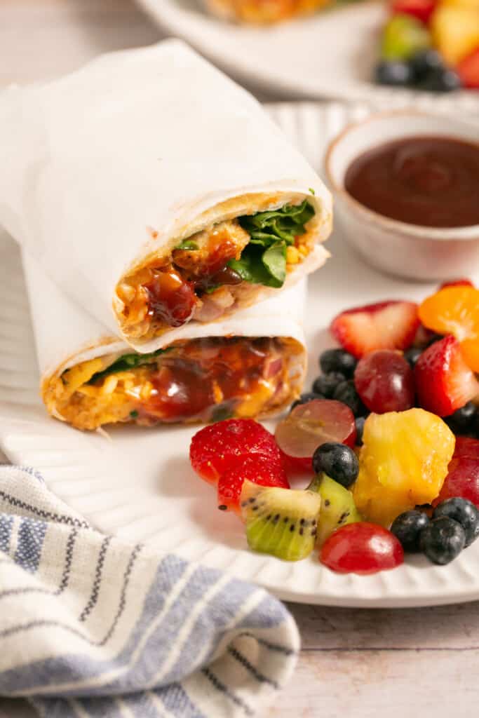 BBQ chicken wrap recipe cut in half stacked vertically on a plate with a side of BBQ sauce and fruit.