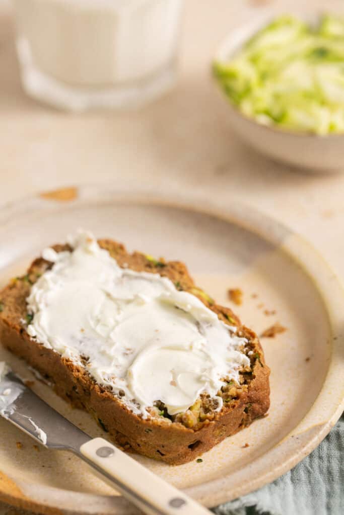 A slice of healthy zucchini bread topped with cream cheese on a plate with a knife.