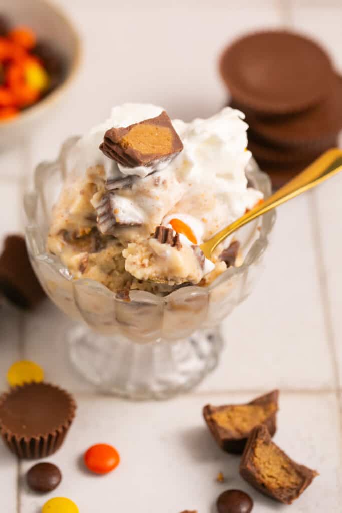Reese's ninja creami protein ice cream in a glass bowl with a spoon topped with whipped cream, Reese's pieces, and more Reese's cups.