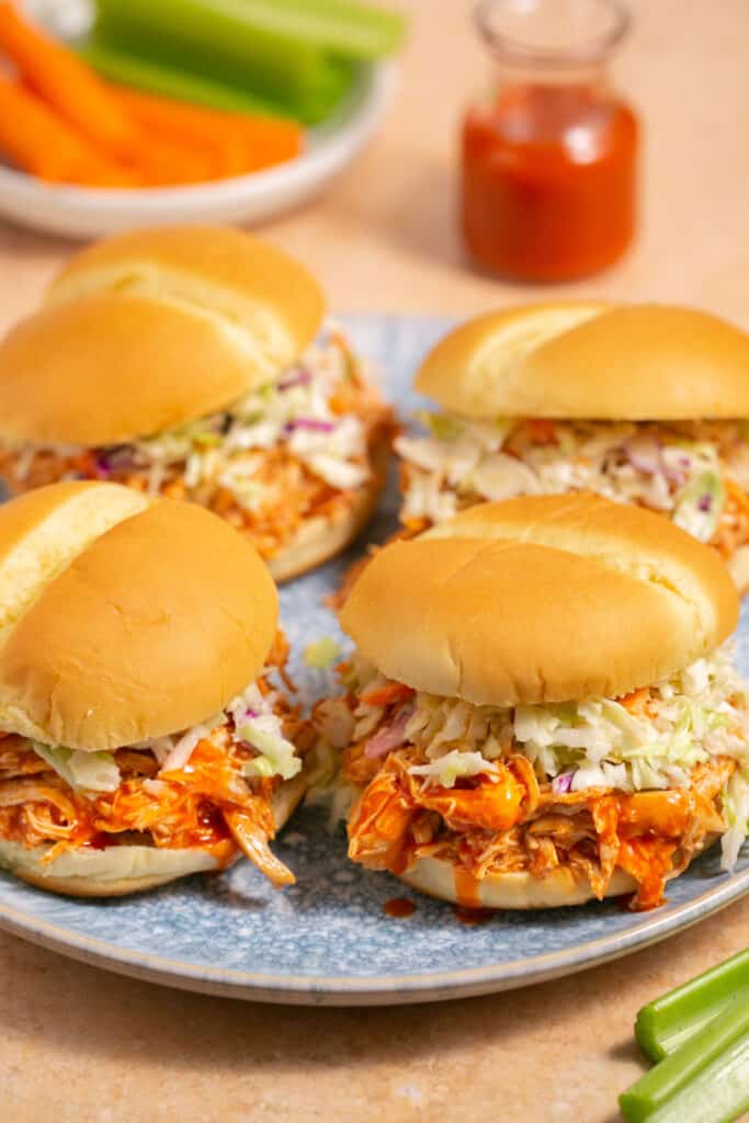 Several pulled buffalo chicken sandwiches topped with coleslaw on buns on plate. 
