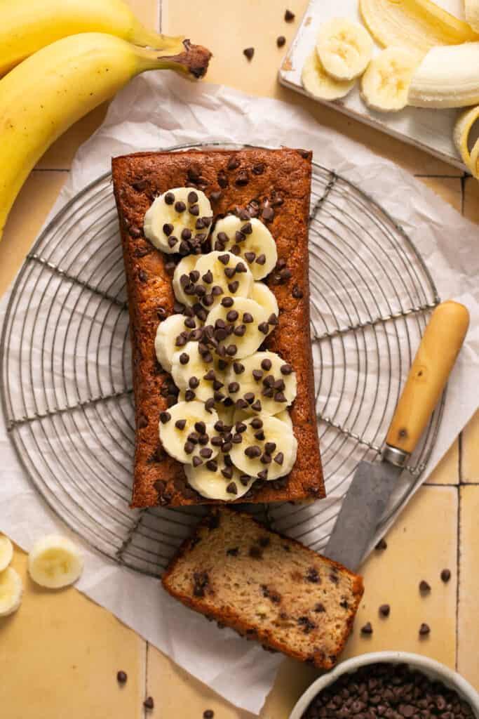 Skinny chocolate chip banana bread topped with banana slices and chocolate chips on a cooling rack.