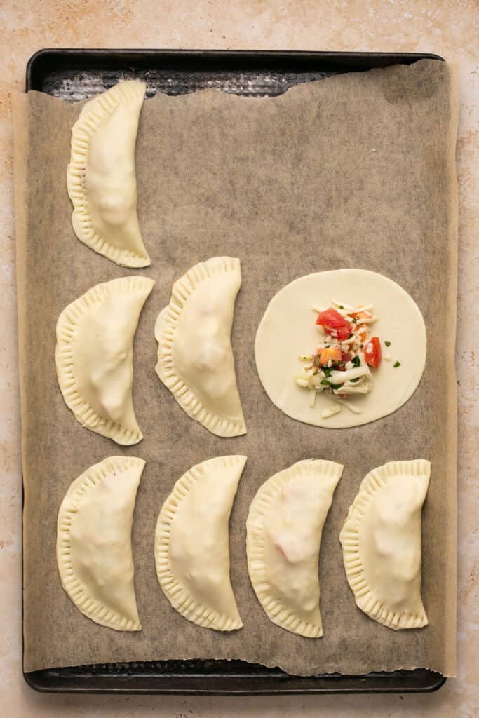 Caprese empanadas on a baking sheet being filled with filling and folded.