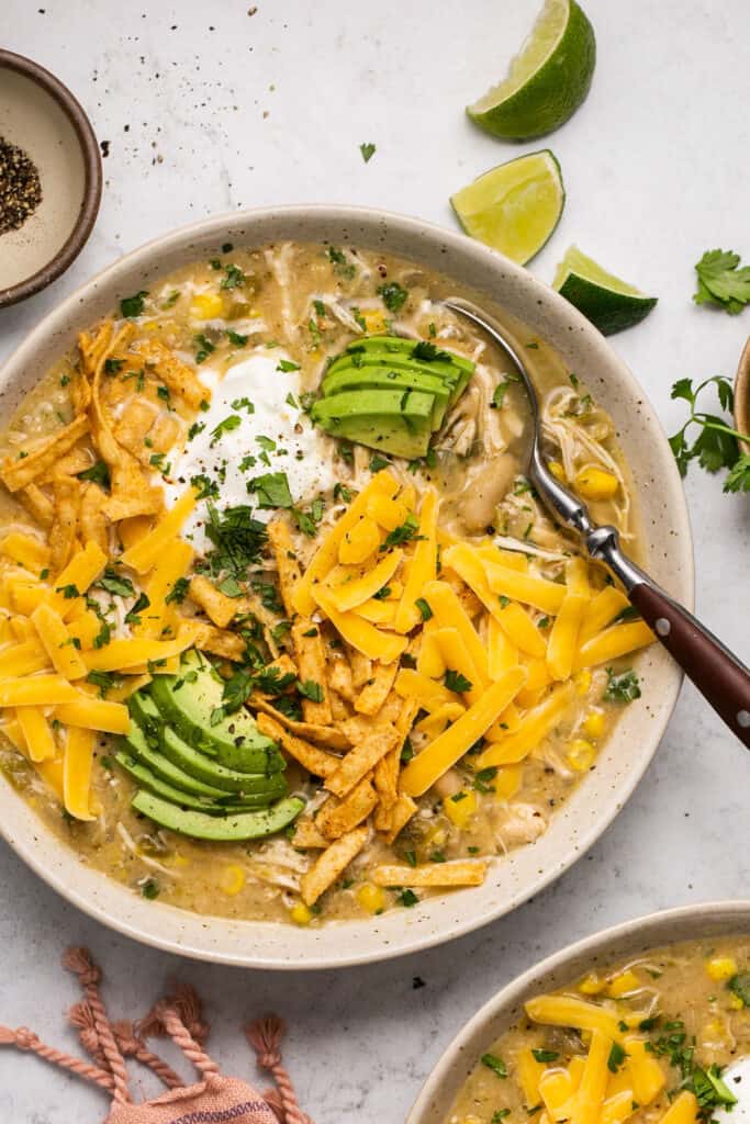 Healthy Crockpot White Chicken Chili topped with shredded cheese, avocado, and sour cream in a bowl with a spoon.