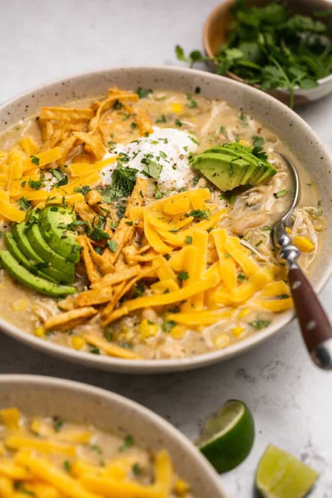 Zoomed in view of Healthy Crockpot White Chicken Chili topped with shredded cheese, avocado, and sour cream in a bowl with a spoon.