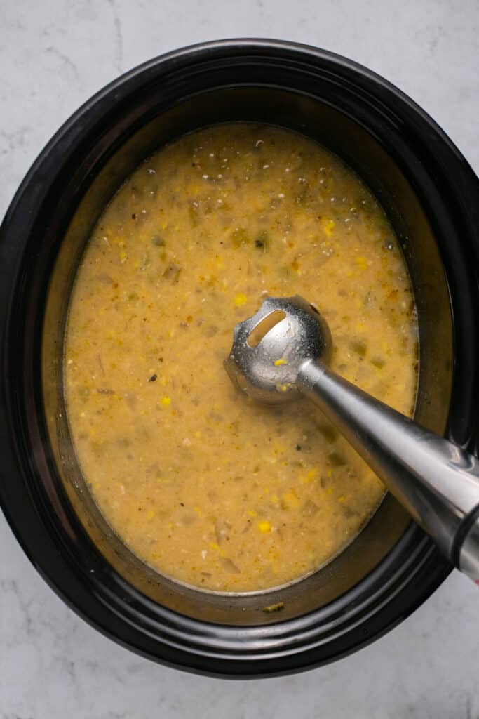 An immersion blender being used in the Healthy Crockpot White Chicken Chili in the slow cooker.