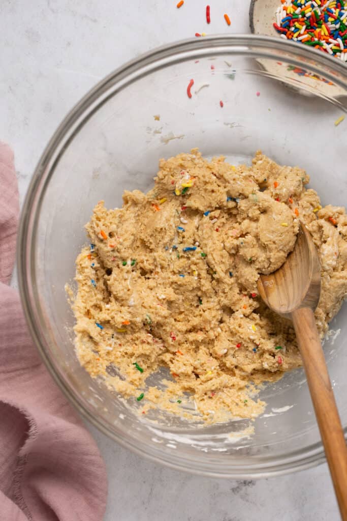 Funfetti protein cookie dough in a mixing bowl with a wooden spoon.