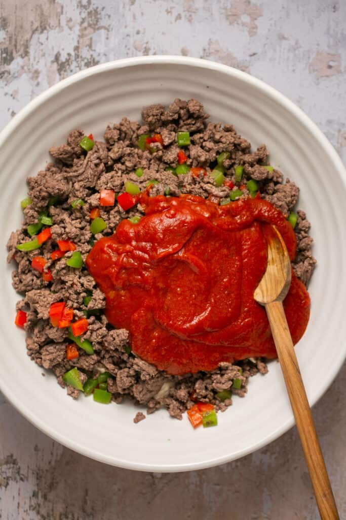 Ground beef and peppers being mixed with pizza sauce in a bowl with a wooden spoon.