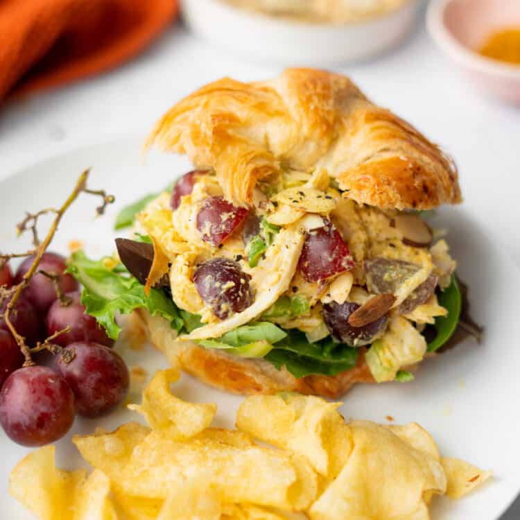 Healthy curry chicken salad on a croissant with grapes and chips on a plate.