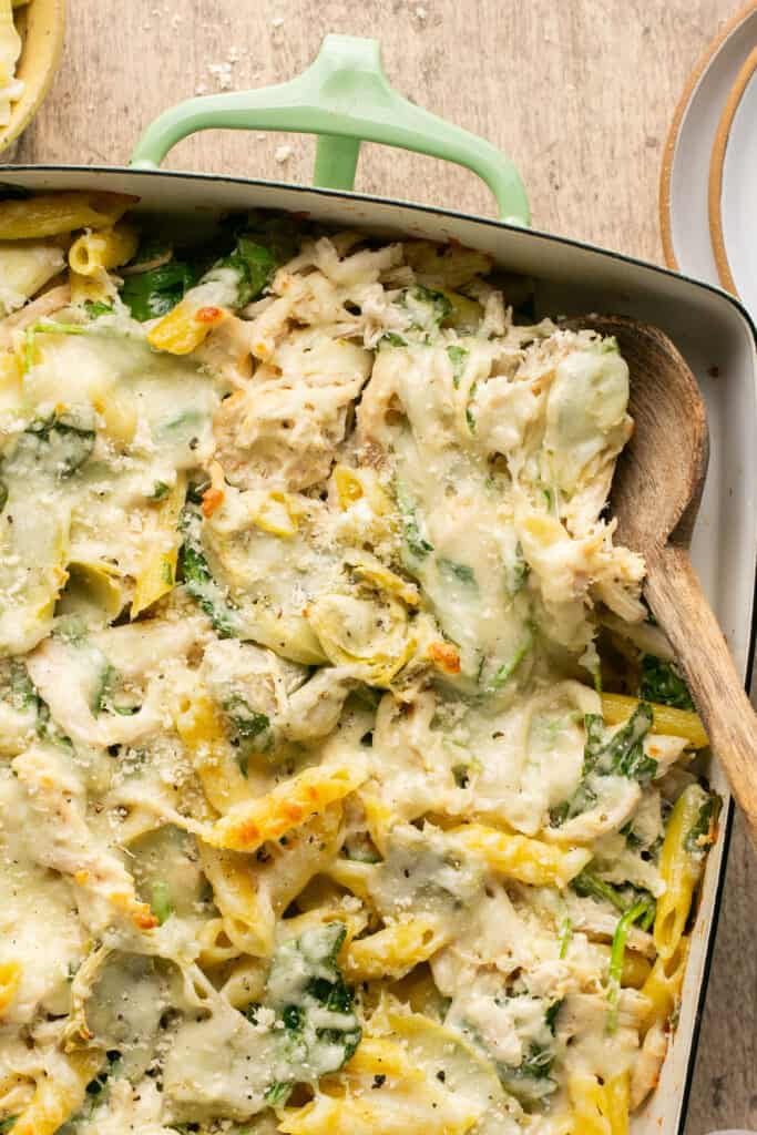 Zoomed in view of healthy spinach and artichoke pasta bake in a baking dish with a wooden spoon.