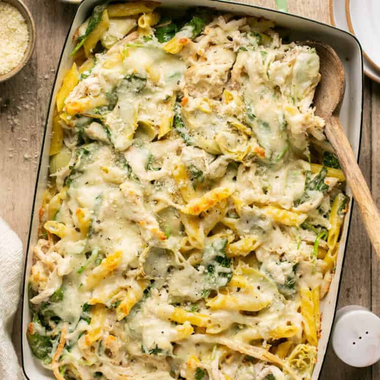 Healthy spinach and artichoke pasta bake in a baking dish with a wooden spoon.