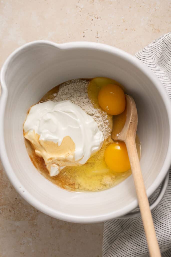 Yogurt and eggs added to the dry ingredients in a mixing bowl with a wooden spoon.