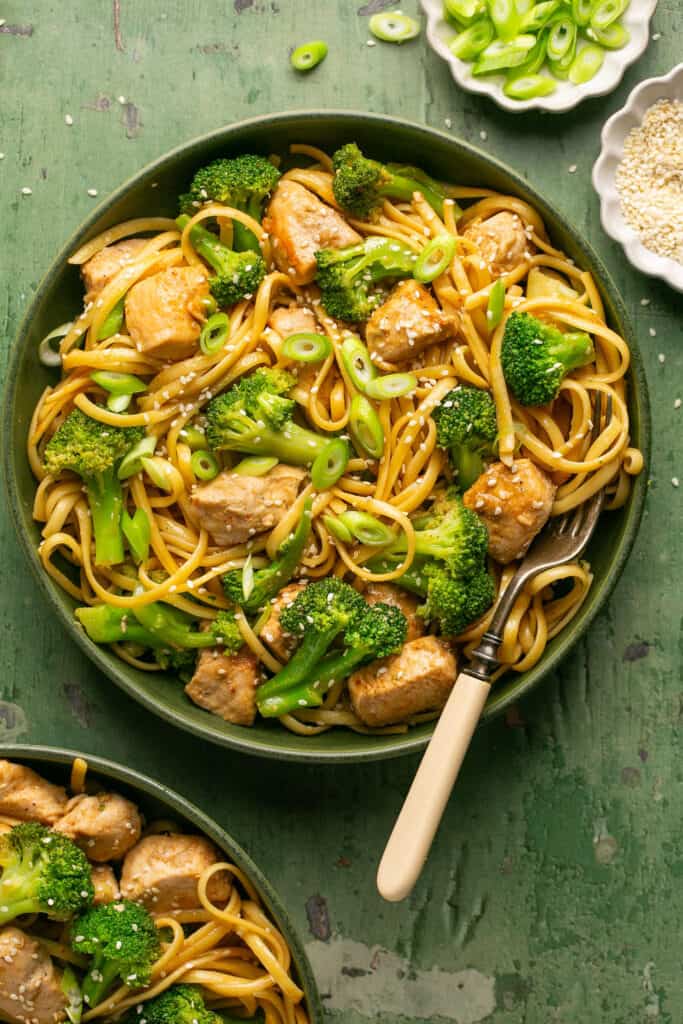 Sesame noodles with chicken and broccoli topped with chopped green onions in a bowl with a fork.