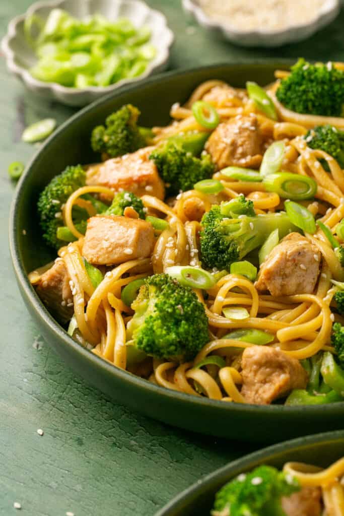 Sesame noodles with chicken and broccoli topped with chopped green onions in a bowl.