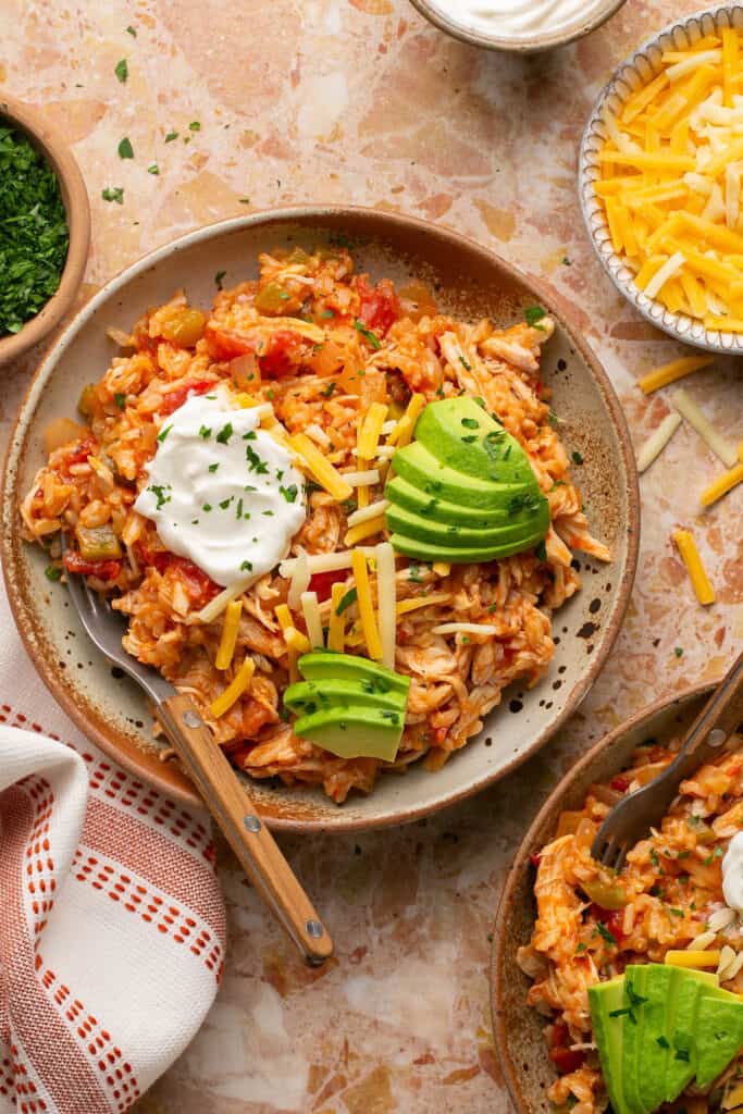 Spanish chicken and rice topped iwth avocado, sour cream, and shredded cheese on a plate with a fork.