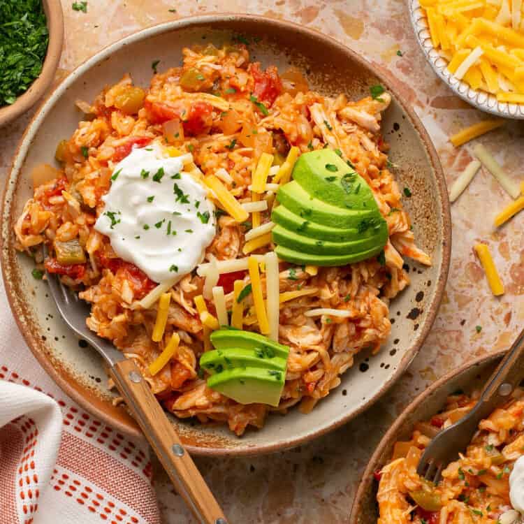 Spanish chicken and rice topped iwth avocado, sour cream, and shredded cheese on a plate with a fork.