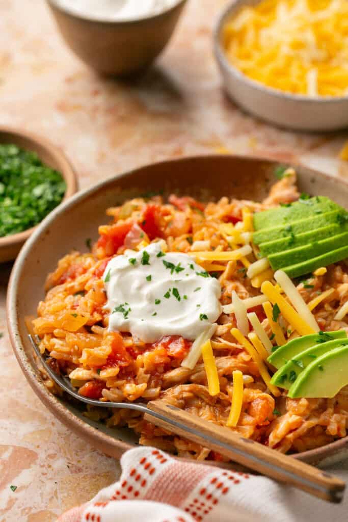 Spanish chicken and rice topped with avocado, sour cream, and shredded cheese on a plate with a fork.