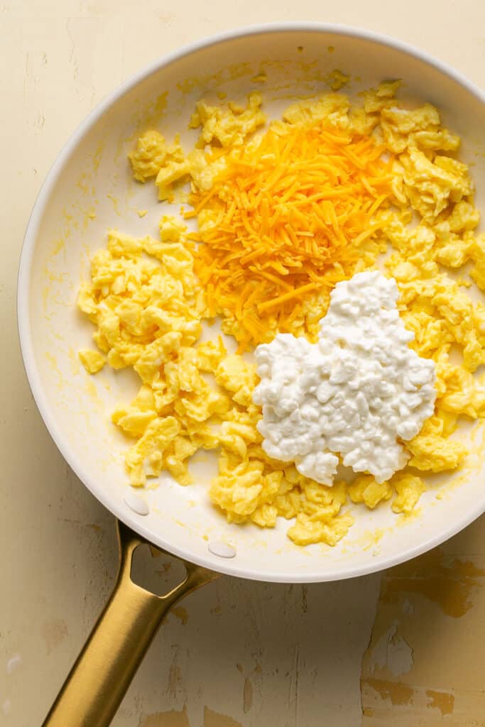 Scrambled eggs in a skillet topped with shredded cheese and cottage cheese before being mixed together.