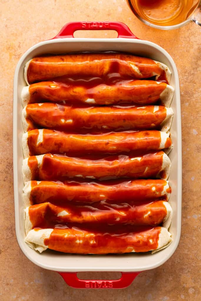 Wrapped tortillas topped with enchilada sauce in a baking dish.