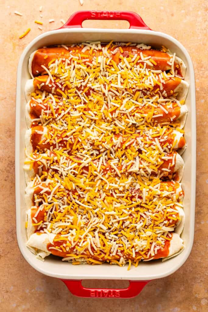 High protein chicken enchiladas topped with shredded cheese in a baking dish before being baked.