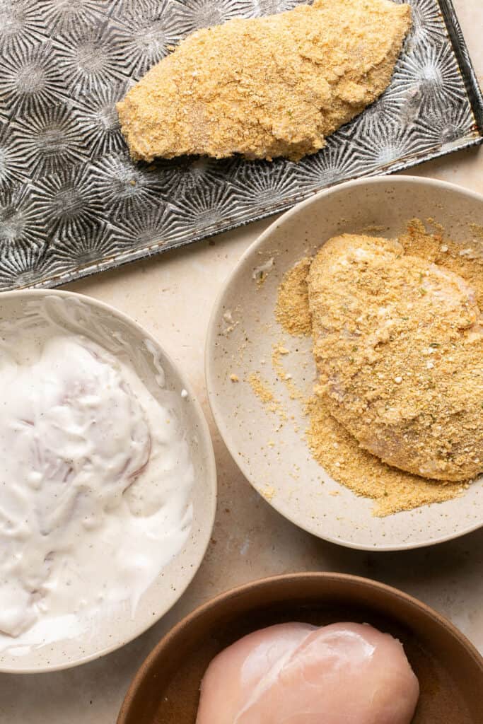 Raw chicken breasts being coated with breadcrumbs.