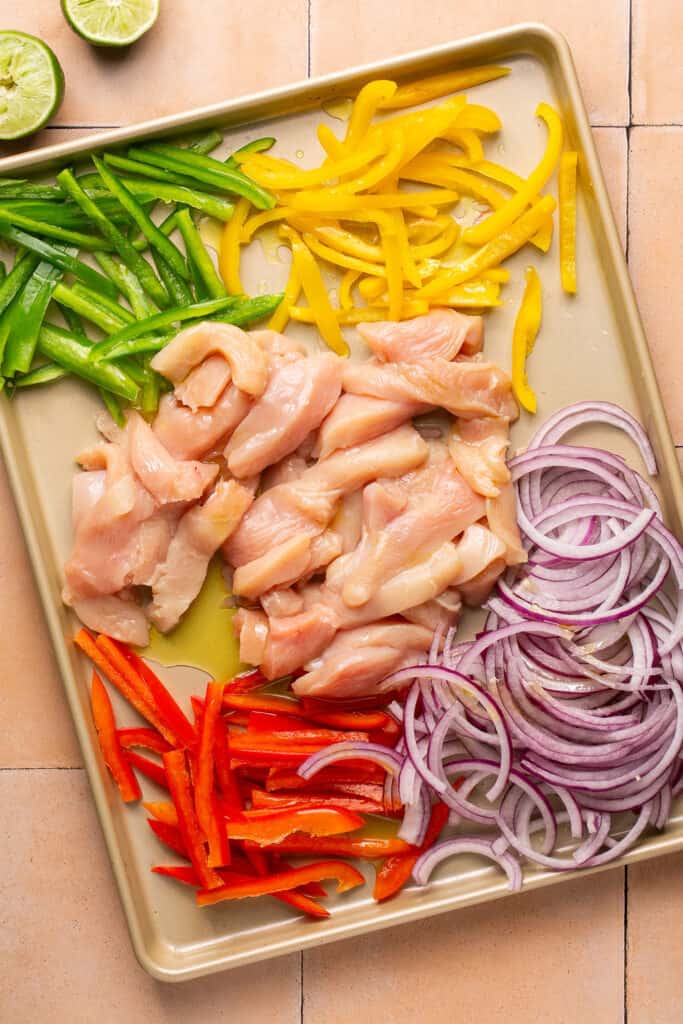 Raw pieces of chicken, slices bell peppers, and red onion on a sheet pan.