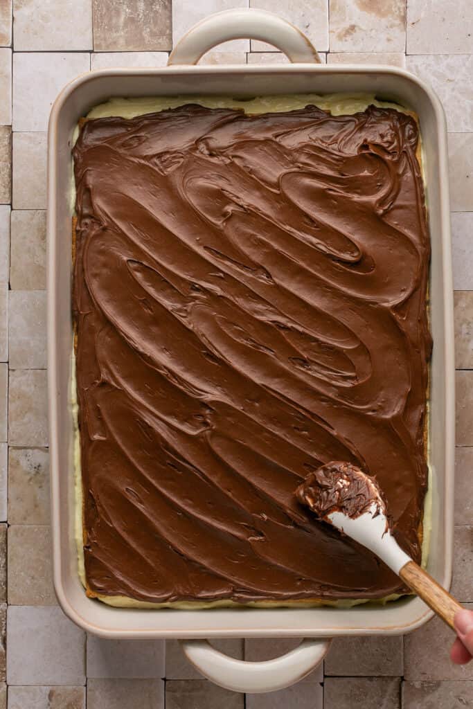 Chocolate frosting being spread on top of the graham cracker layer in the baking dish.