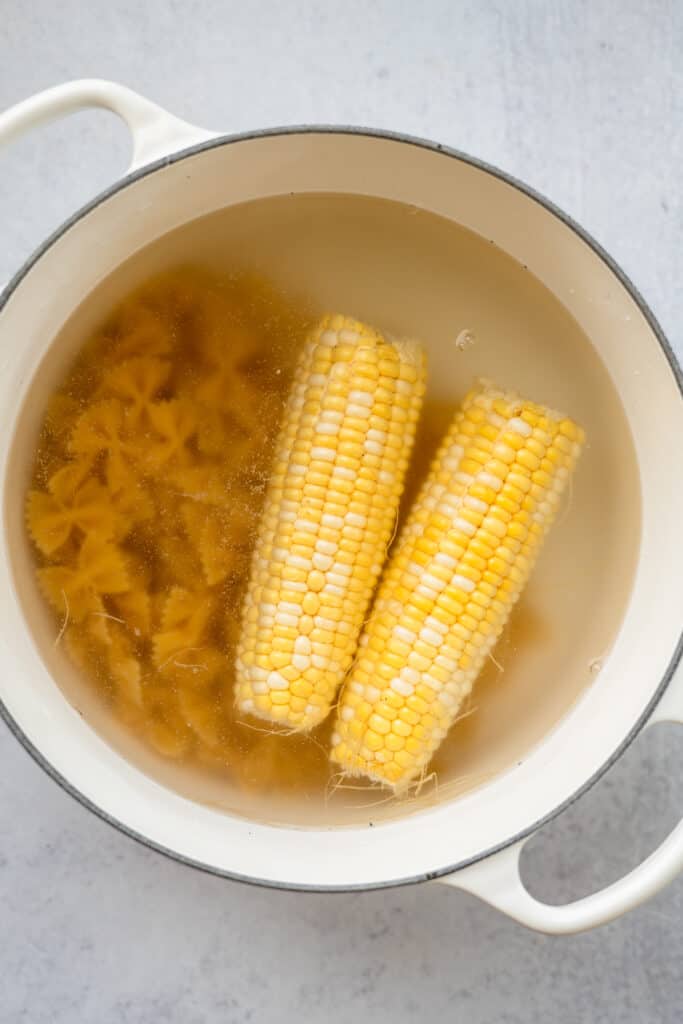 Uncooked pasta and corn on the cob in a pot with water.