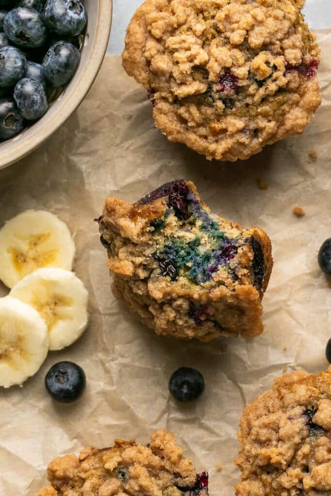 Banana blueberry oatmeal muffins, some cut in half, on parchment paper garished with banana slices and blueberries.
