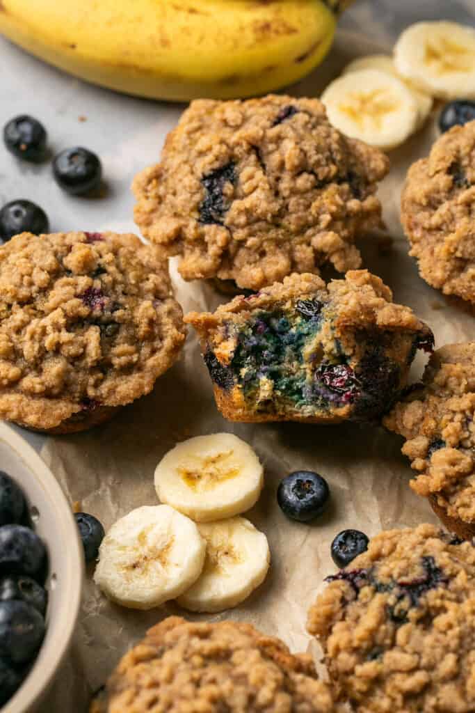 Banana blueberry oatmeal muffins, some cut in half, on parchment paper garnished with banana slices and blueberries.