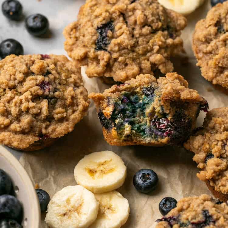 Banana blueberry oatmeal muffins, some cut in half, on parchment paper garnished with banana slices and blueberries.