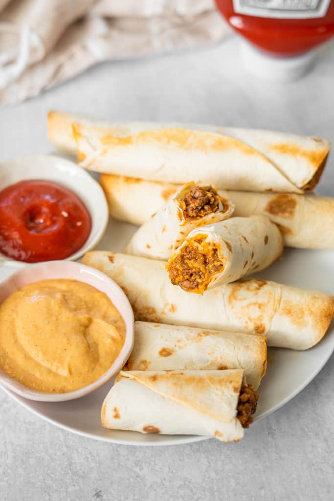 Cheeseburger taquitos on a plate with ketchup and mustard on the side.