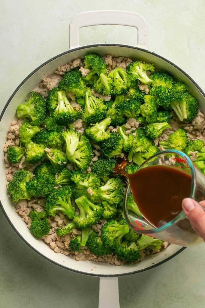 Sauce being poured over the chopped broccoli and cooked ground chicken in a skillet.