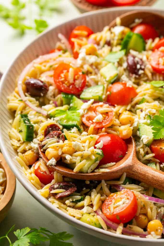 Mediterranean chickpea and orzo pasta salad in a large bowl with a wooden spoon.