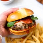 A hand holding a pizza burger with fries on a plate in the background.
