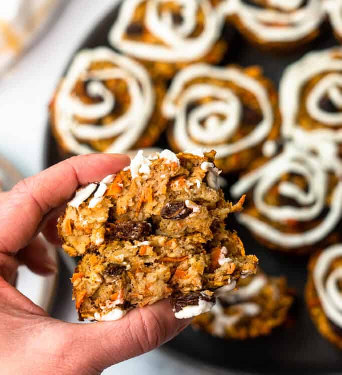 A hand holding a Carrot Cake Baked Oat Cup with a tray of Carrot Cake Baked Oat Cups in the background.