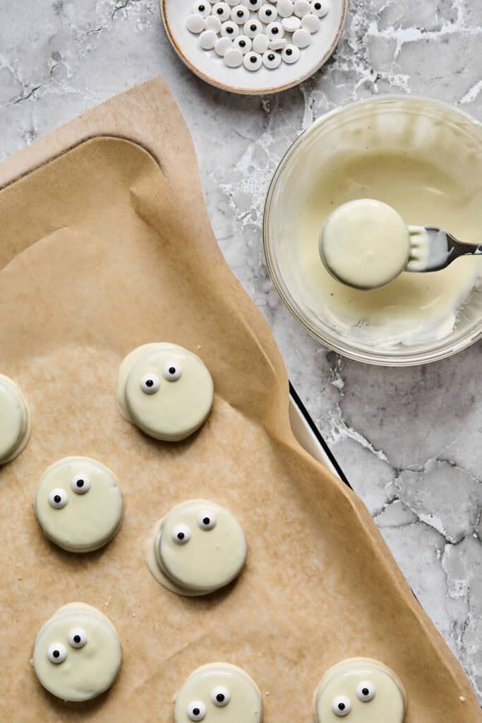 Oreos being dipped in melted white chocolate with a fork and placed on a baking sheet with parchment paper.