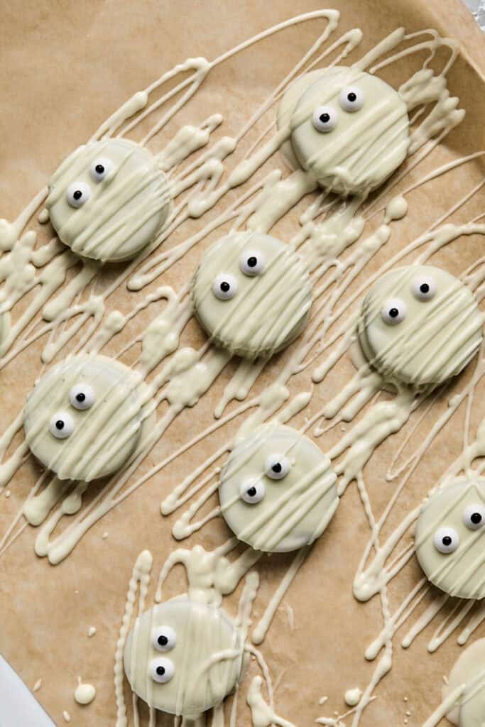 Oreo Mummy Cookies being drizzled with white chocolate on a baking sheet with parchment paper.