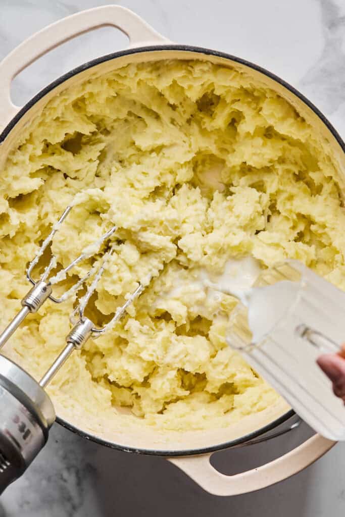 Milk being added to the potato mixture being mixed with an electric mixture in a pot.