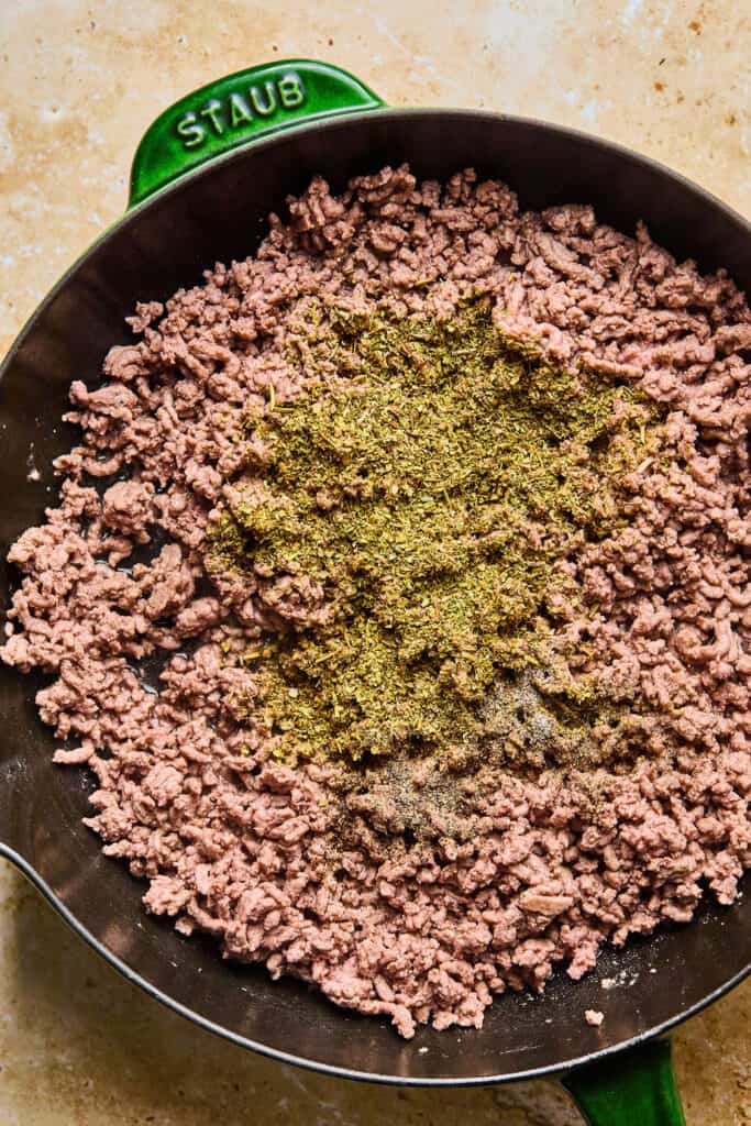 Cooked ground beef topped with seasonings in a skillet.