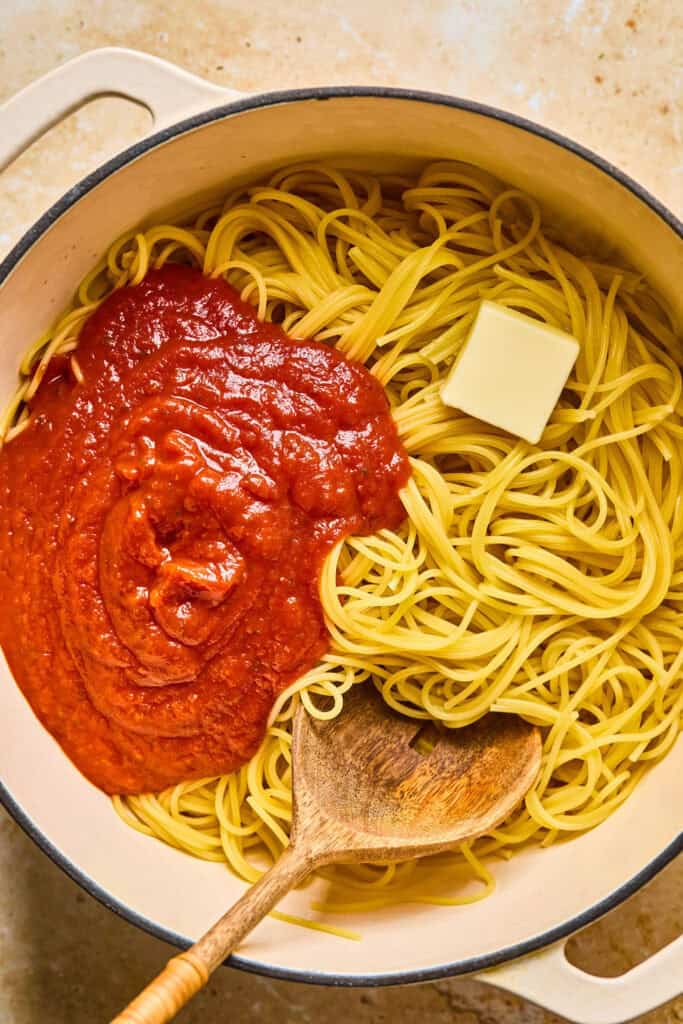 Cooked spaghrtti, tomato sauce, and butter in a bowl with a wooden spoon.