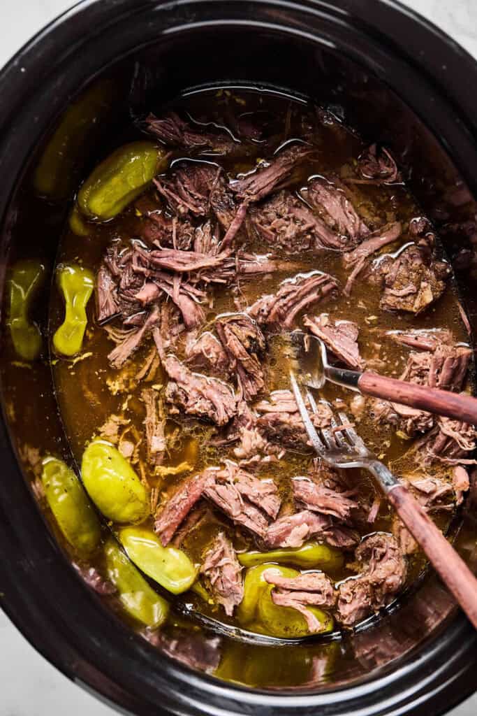 Mississippi Pot Roast being shredded with two forks in a slow cooker.