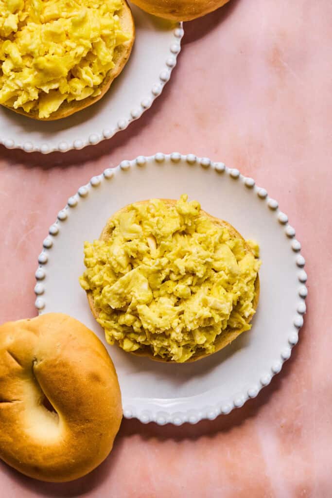 Scrambled eggs on a small plate with a bagel on the side.